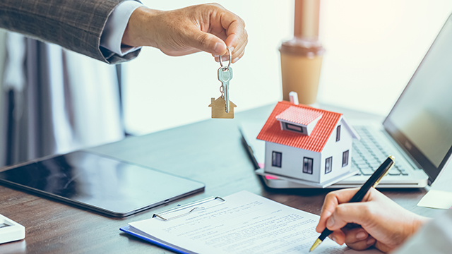 close person handing over the keys to a new home to another person signing mortgage documents at a table.